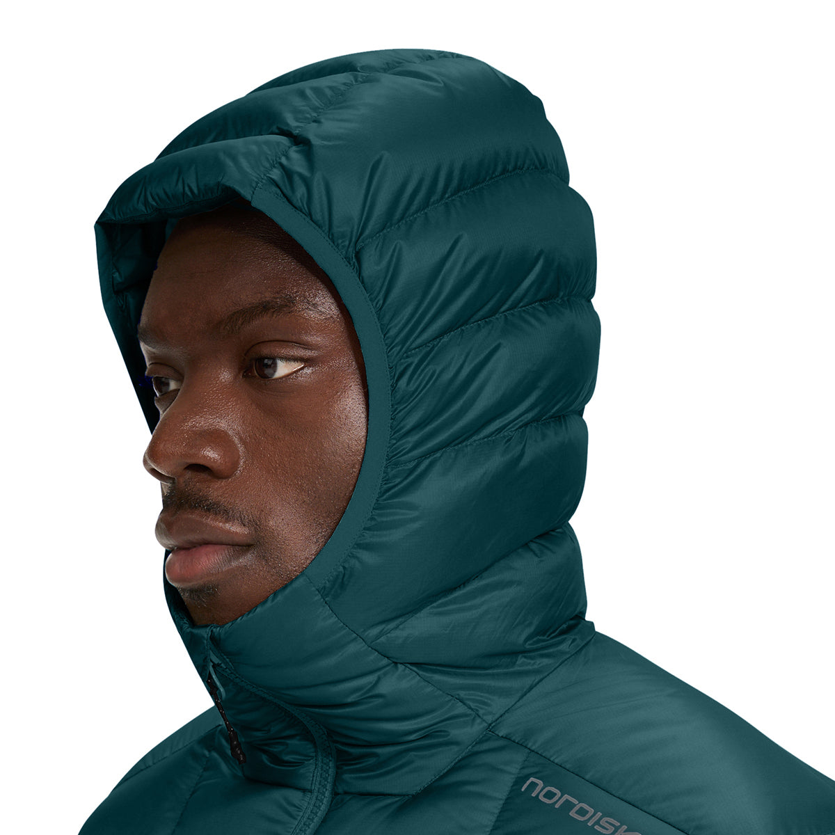 A close-up side profile of a man wearing a dark teal hooded puffer jacket against a solid white background. The brand name 'nordisk' is visible on the lower right chest of the jacket.