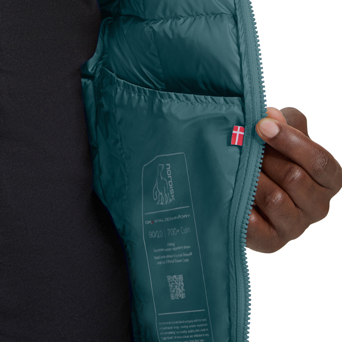 Close-up of a dark teal puffer jacket interior pocket with a hand holding the zipper, showing printed label details and a small red-and-white flag tag.