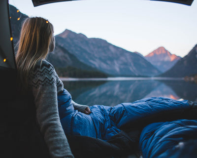 Woman in a blue summer sleeping bag looking at a mountain lake from a tent.