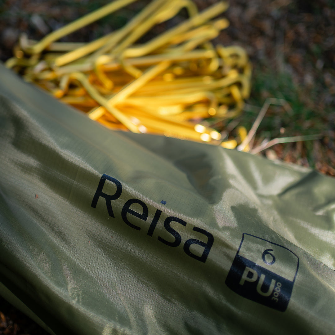 A close-up of an olive green tent fabric with the brand name 'Reisa' and a 'PU 3000' label printed on it, with yellow tent stakes blurred in the background on a forest floor.