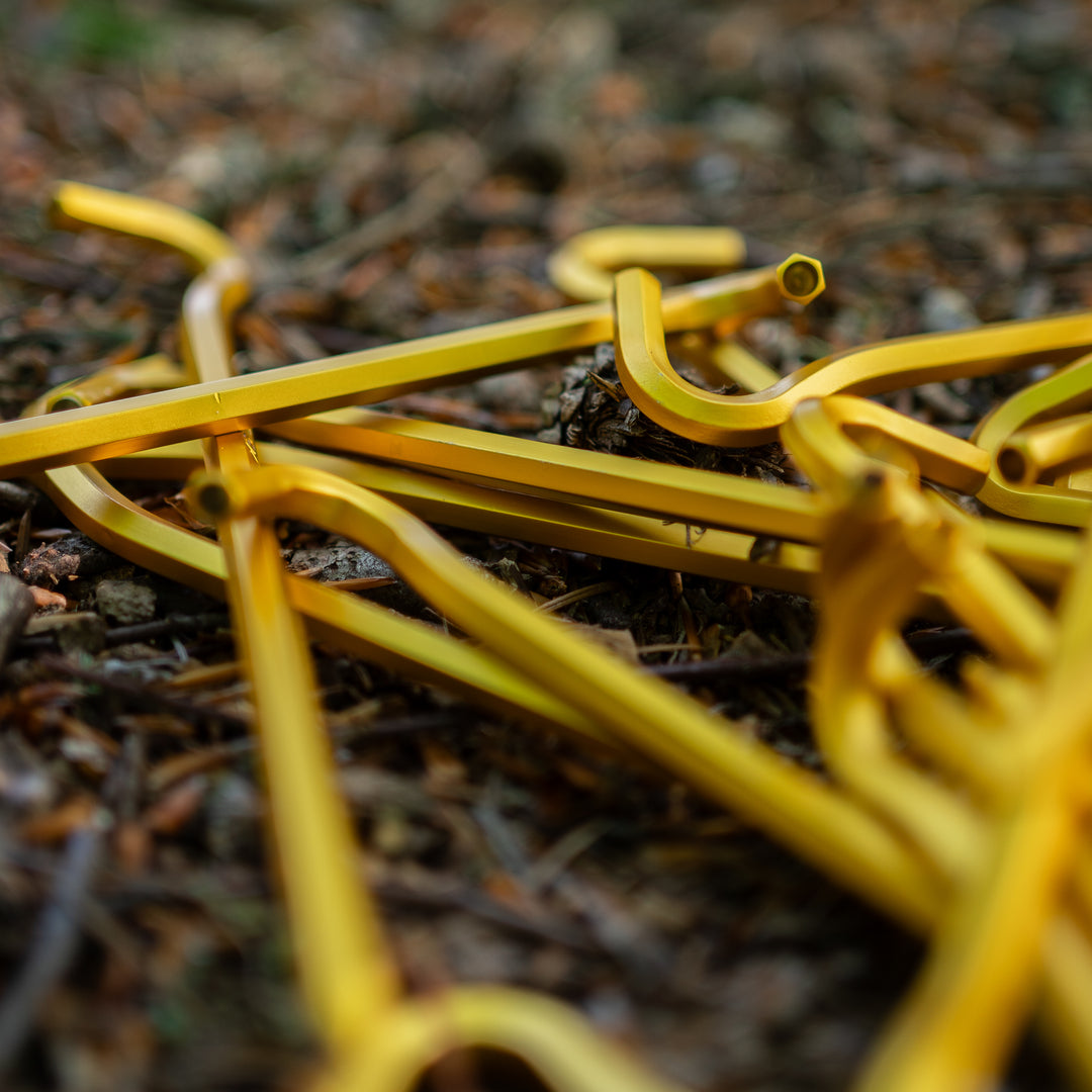 A pile of bright yellow metal tent stakes or guying rods lying on a forest floor covered in pine needles and leaves.