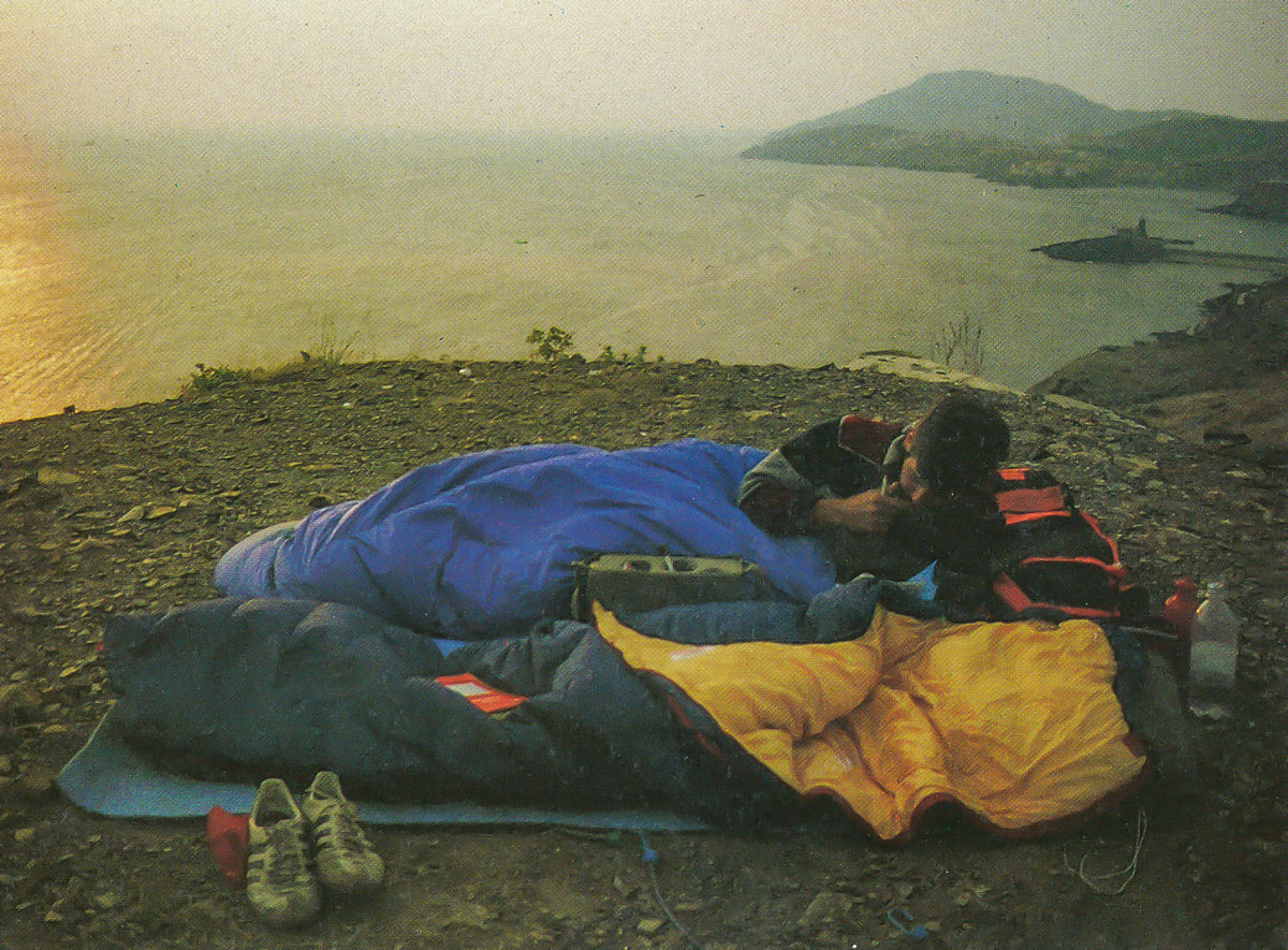 Vintage photo of person camping outdoors in sleeping bags on a rocky cliff overlooking the ocean.