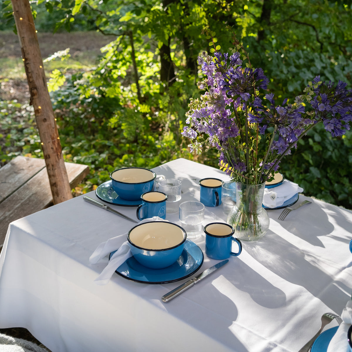 Blue Nordisk Madam Blå enamel cups and bowls set on a white outdoor table.