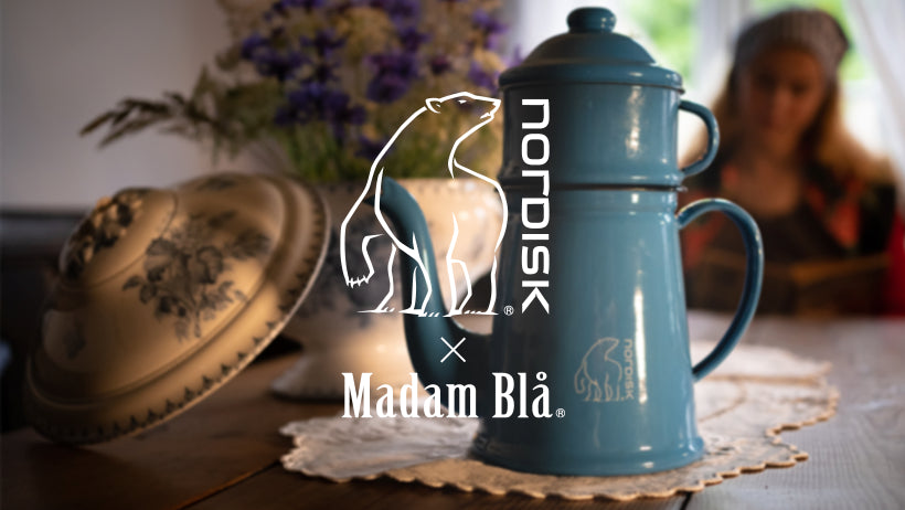 Blue Nordic enamel coffee pot with Madam Blå and Nordisk logo on a wooden table.