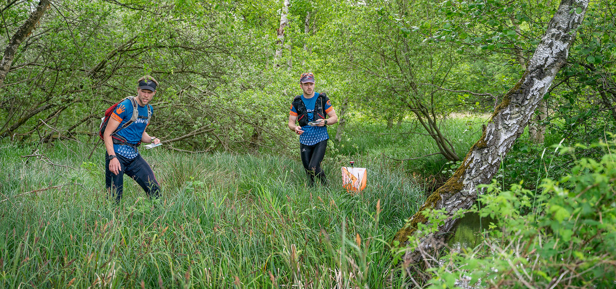 Two runners participating in the Nordisk Adventure Run Silkeborg orienteering through a lush green forest.