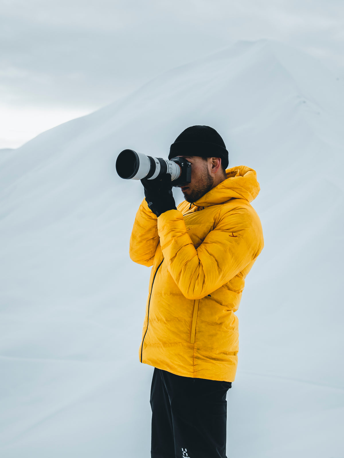 Nordisk Hero Rico taking photos in a yellow down jacket against a snowy mountain background.