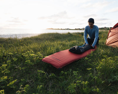 Man rolling up a sleeping bag on a red Nordisk sleeping mat at a campsite.