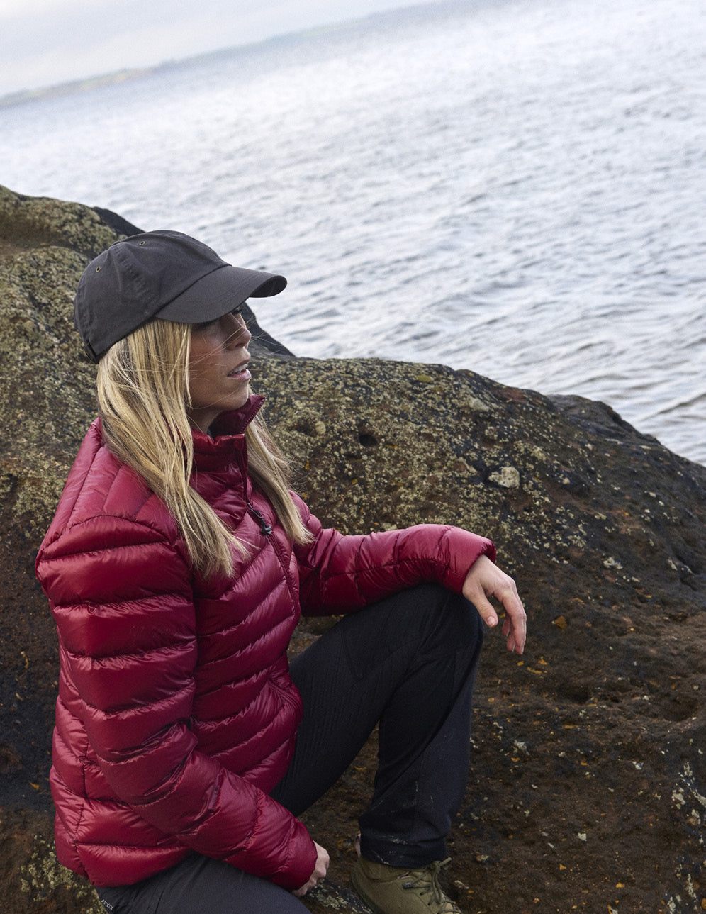 Woman wearing the Companyon Thyra down jacket in Tibetan Red, sitting on a rock by the coast