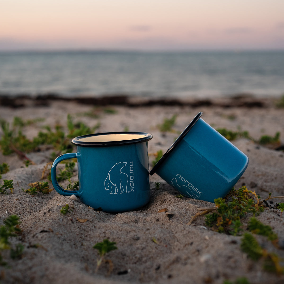 Two blue Madam Blå enamel mugs with Nordisk logo on a sandy beach at sunset.