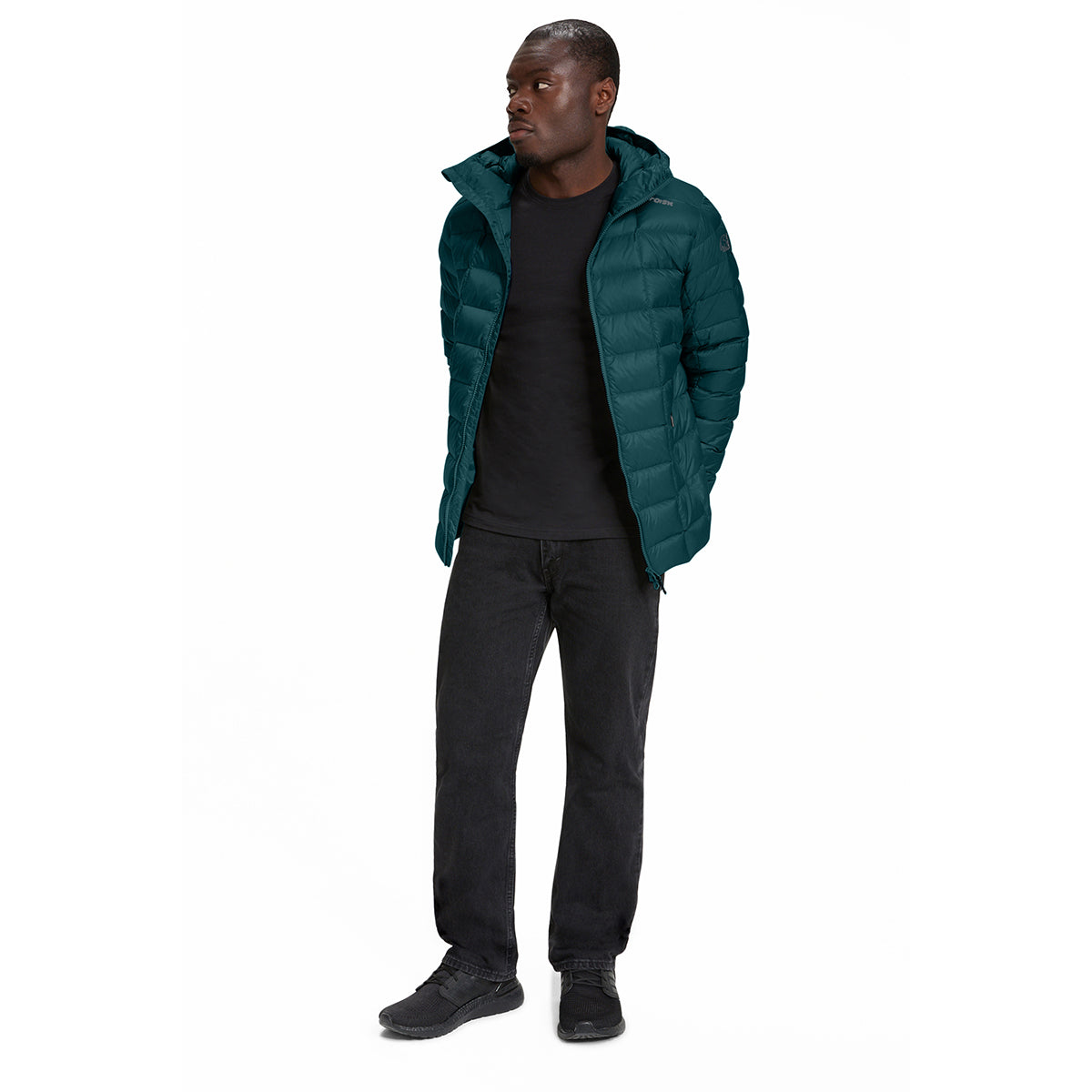 A man wearing a dark teal-green puffer jacket over a black t-shirt, black jeans, and black sneakers, standing against a solid white background.