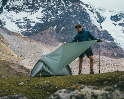 Hiker setting up a green Nordisk lightweight tarp over a tent in snowy mountain terrain.