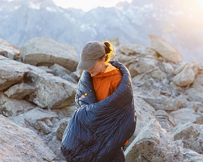 Woman wrapped in a navy Nordisk sleeping bag on a rocky mountain landscape at sunrise.