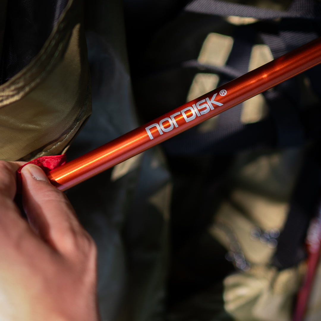 A close-up shot of a person's hand holding an orange-red trekking pole with the brand name 'nordisk' printed in white on the aluminum shaft. The background is a dark, out-of-focus outdoor gear bag.