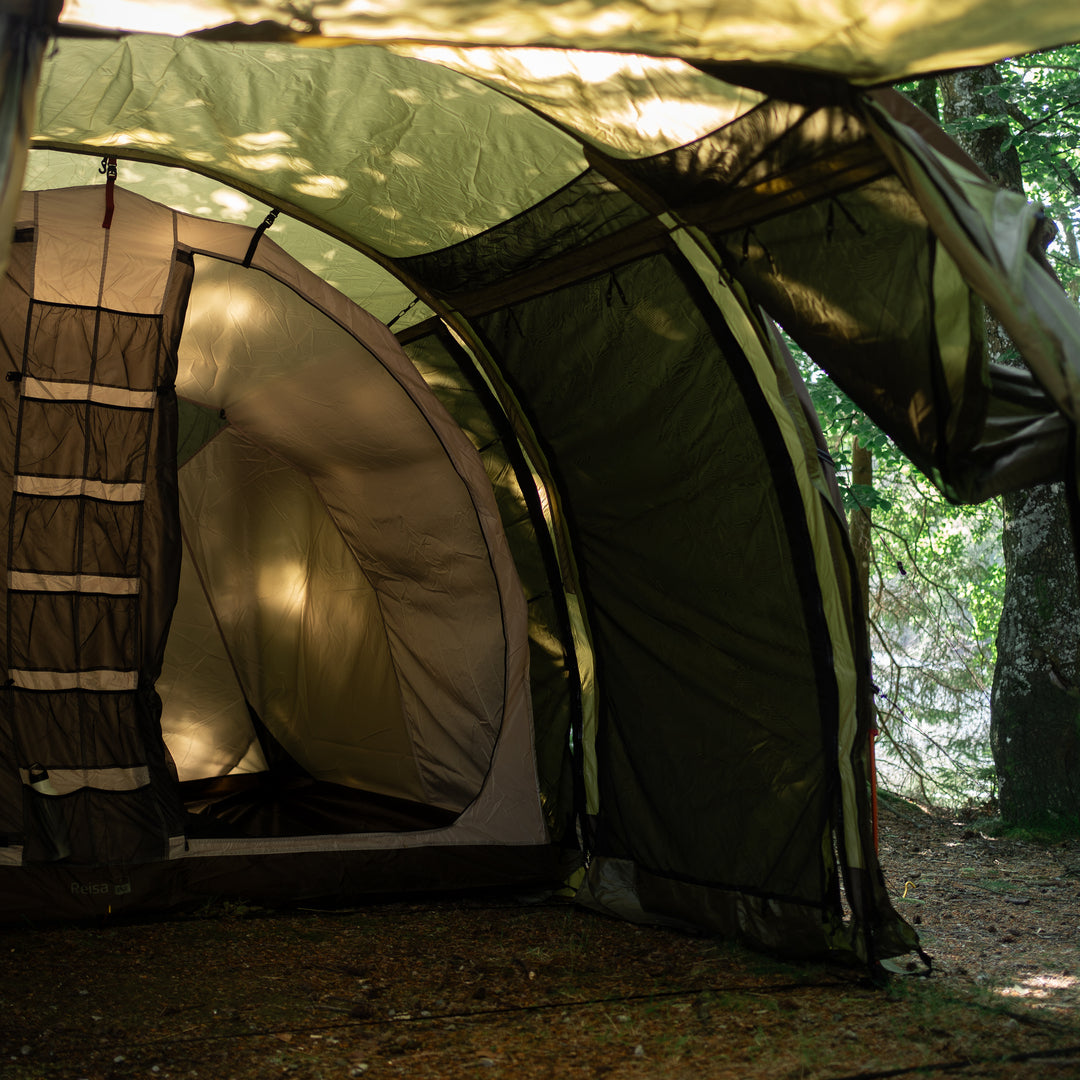 A dome-style camping tent pitched outdoors in a wooded setting, with its front door open and sunlight filtering through the fabric and surrounding trees.