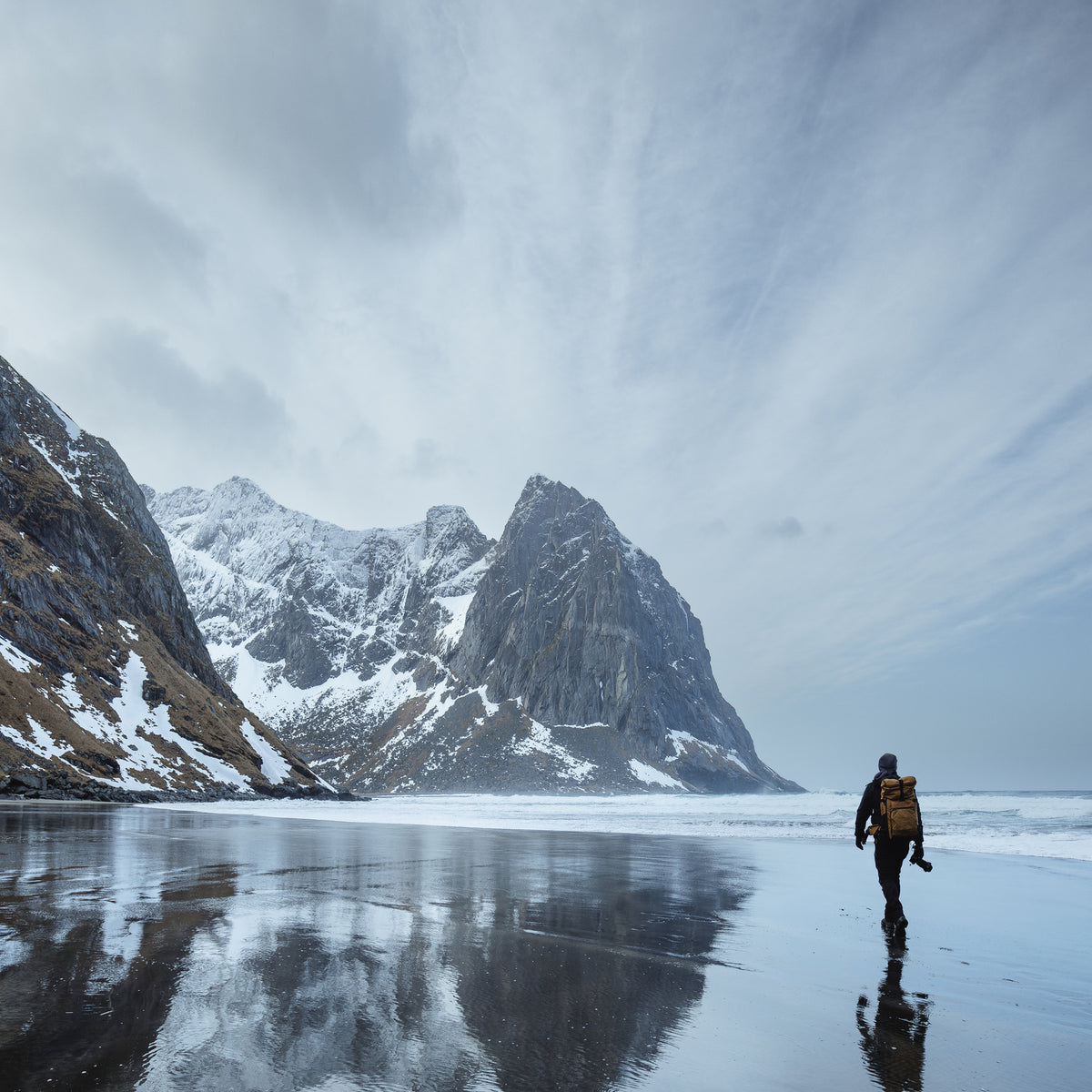 Man hiking on a snowy Arctic beach in Norway with a backpack and camera gear.