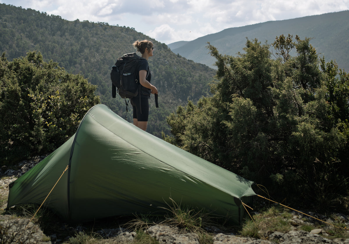 Person with a backpack standing near a green tent in a mountainous area.